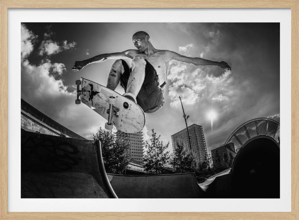 A dramatic low-angle, black and white photograph of a shirtless skateboarder caught in mid-air above a skatepark ramp. His arms are spread wide for balance as the sun flares brightly behind him, set against a cloudy urban sky with high-rise buildings in the distance. The image is enclosed in a silver frame. Wall Art