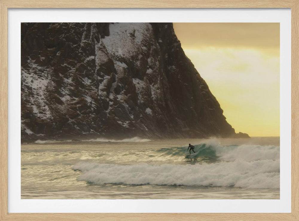 A framed photograph captures a lone surfer in a black wetsuit riding a turquoise wave. In the background, a massive, steep, snow-dusted mountain looms over the cold sea under a pale yellow, overcast sky. Print