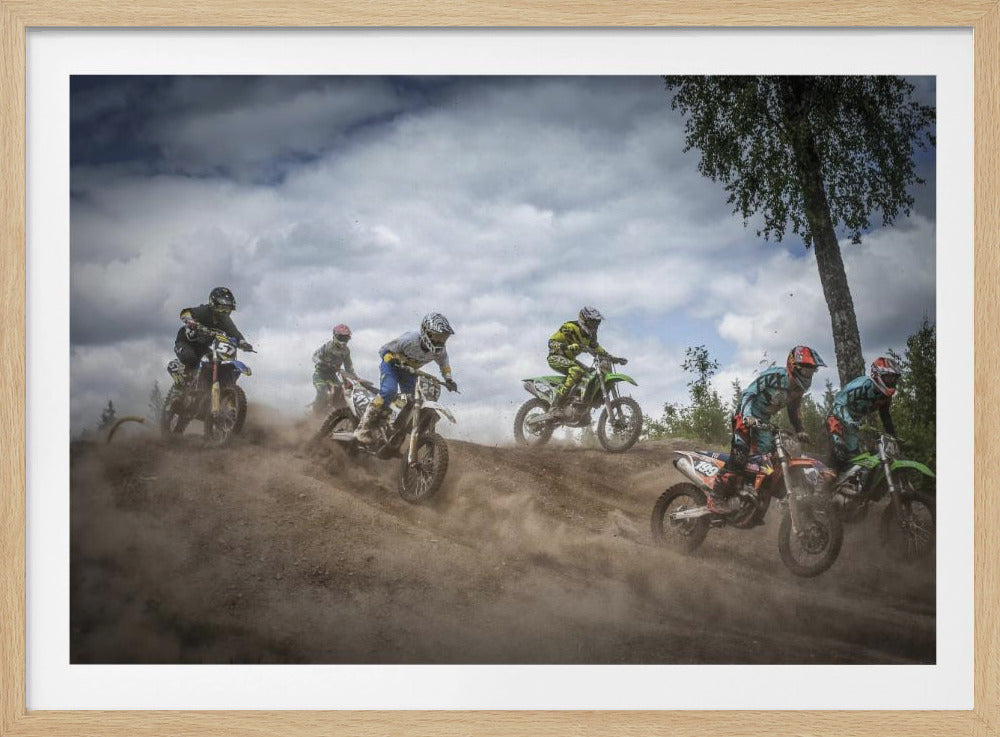 A dynamic action shot of several motocross riders racing up a dirt hill, kicking up a large cloud of dust. The riders are in full gear on colorful bikes, with a dramatic cloudy sky and a tall tree in the background. Print