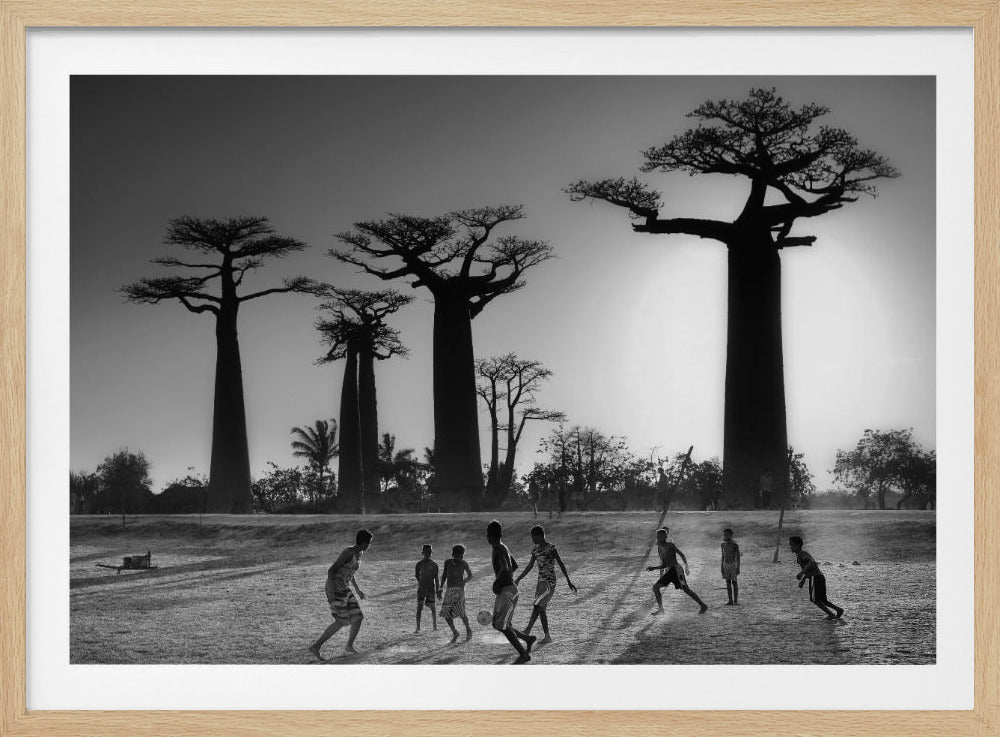 A framed, black and white photograph capturing a vibrant scene of several children playing soccer on a dirt field. In the background, the iconic silhouettes of large Baobab trees stand tall against a bright, hazy sun, casting long shadows of the players across the ground. Artwork
