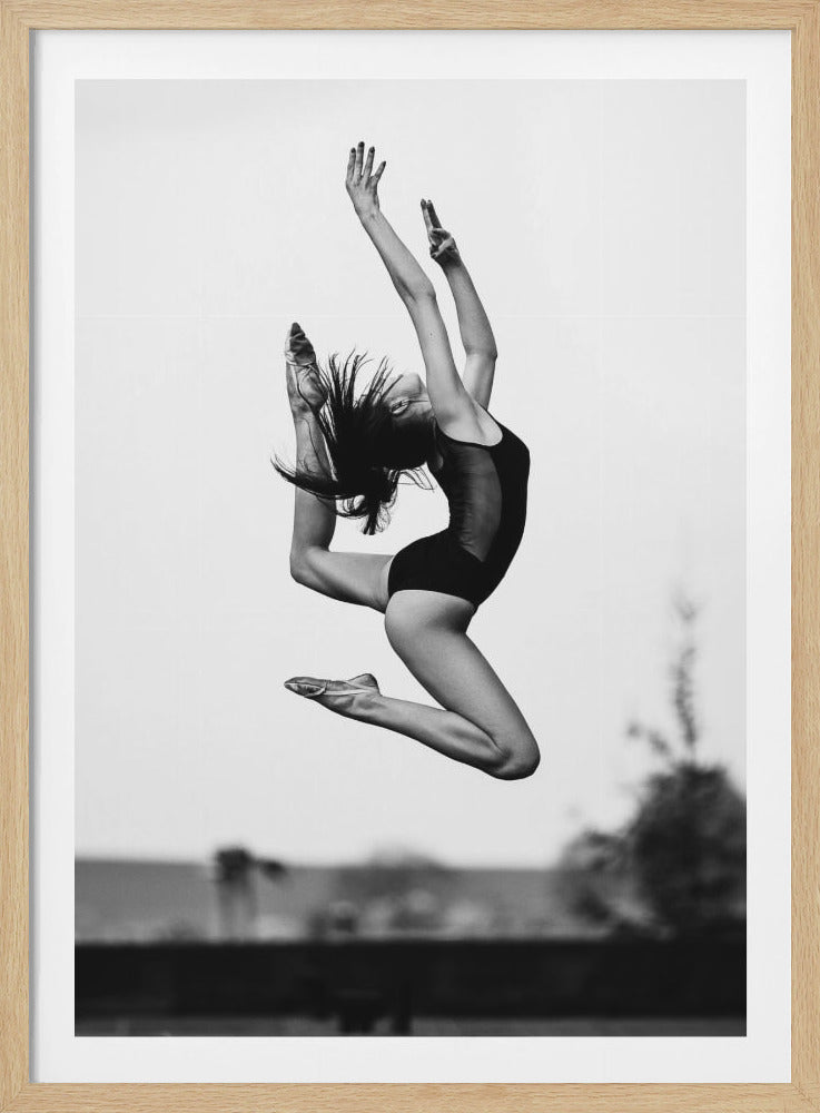 A dynamic black and white photograph of a female ballet dancer caught mid-leap against a light, blurred background. She wears a dark leotard and ballet slippers, her body is arched back in an impressive display of flexibility and grace, with her arms extended upwards and hair flying from the movement. Decor