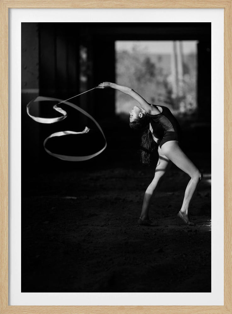 A dramatic black and white photograph of a female rhythmic gymnast in profile. She is arched backward in a dynamic pose inside a dark, shadowed space, twirling a long ribbon that creates graceful curves in the air. Her form is highlighted by a soft light. Poster