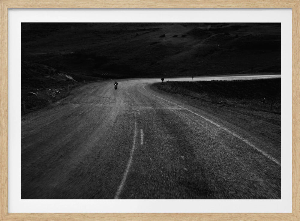 A moody, black and white photograph capturing a lone motorcyclist from behind as they navigate a curving road through a dark, hilly terrain, framed in silver. Decor