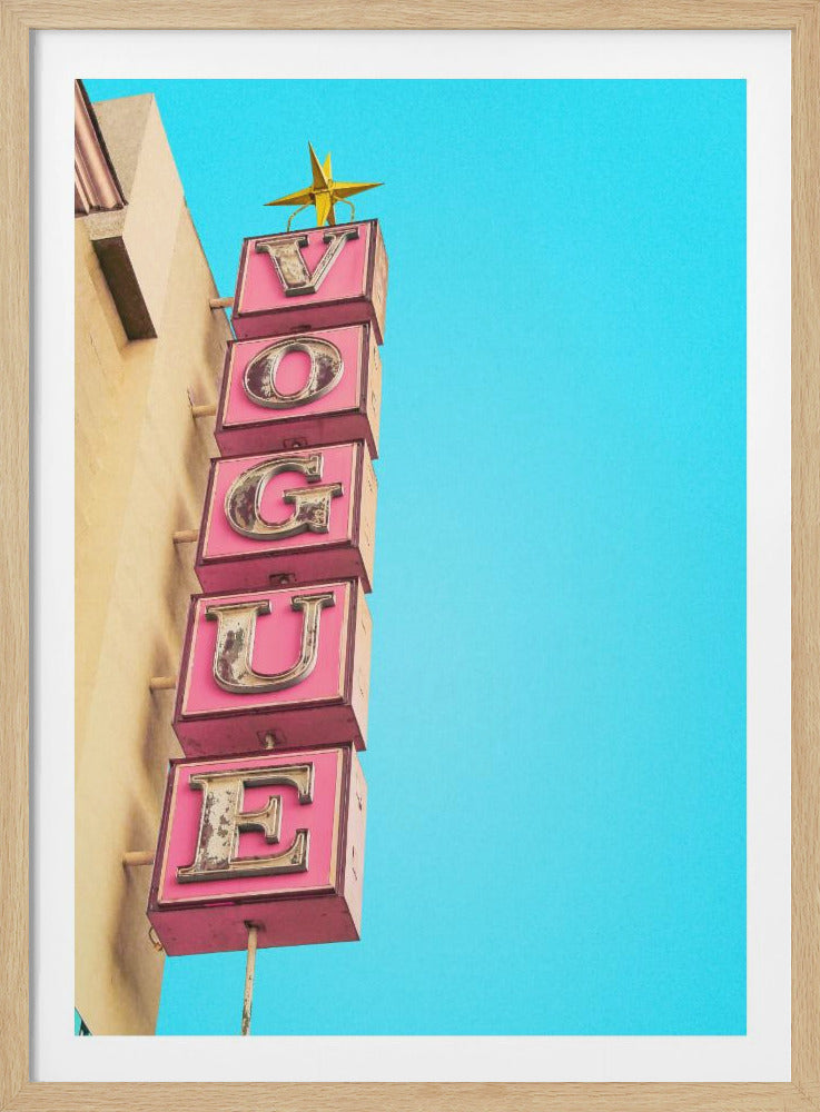 A low-angle shot of a vintage vertical sign spelling 'VOGUE' in pink block letters with a weathered metallic finish, set against a solid bright turquoise sky. The sign is attached to the side of a tan building and is topped with a golden star. Wall Art