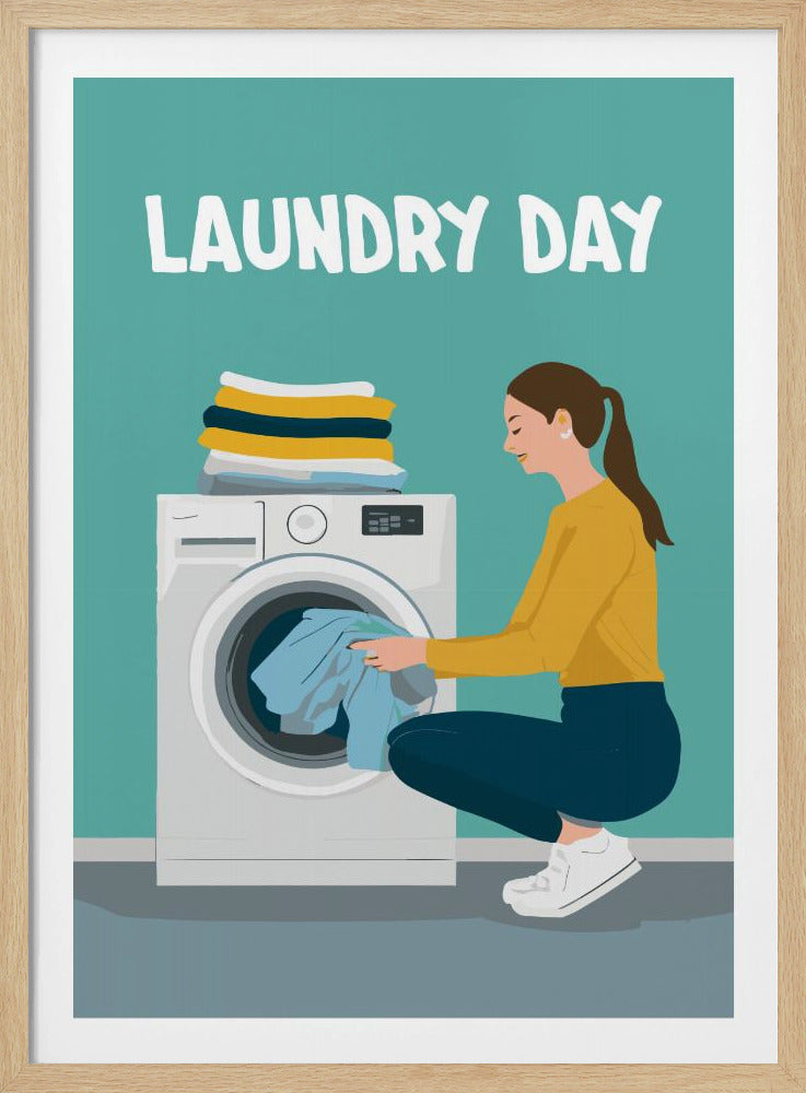 A framed illustration poster with the words 'LAUNDRY DAY' in white at the top. A woman with brown hair in a ponytail, wearing a yellow top and blue pants, crouches to load blue clothes into a white front-loading washing machine. A stack of folded laundry sits atop the machine against a teal background. Print