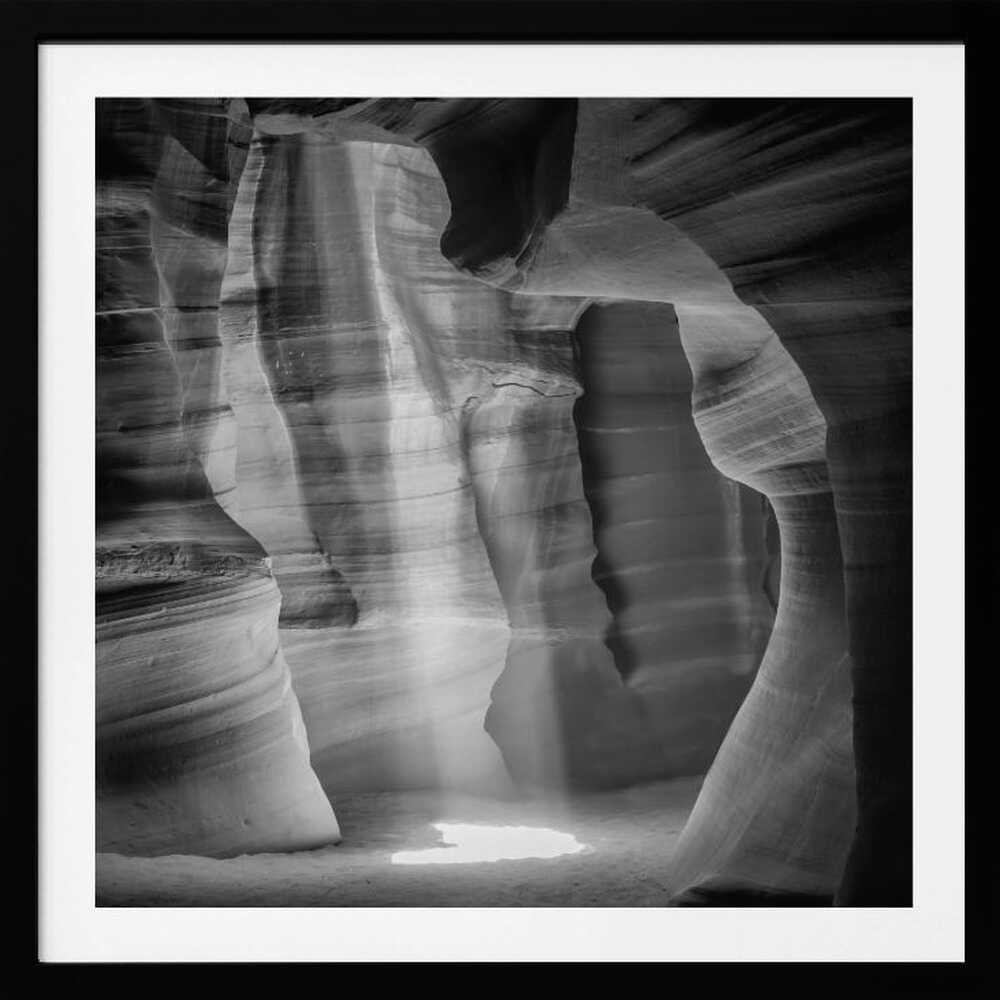 A dramatic black and white photo of a sunbeam lighting the sandy floor of a smooth, curved slot canyon.