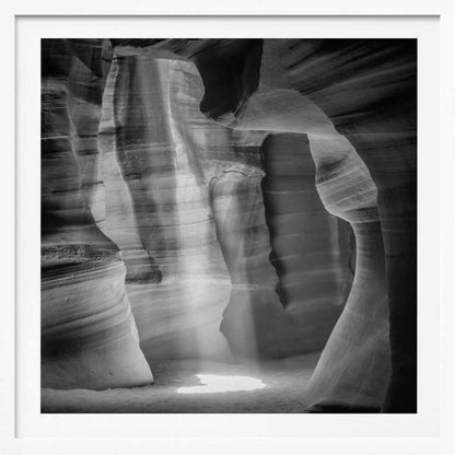 A dramatic black and white photo of a sunbeam lighting the sandy floor of a smooth, curved slot canyon.