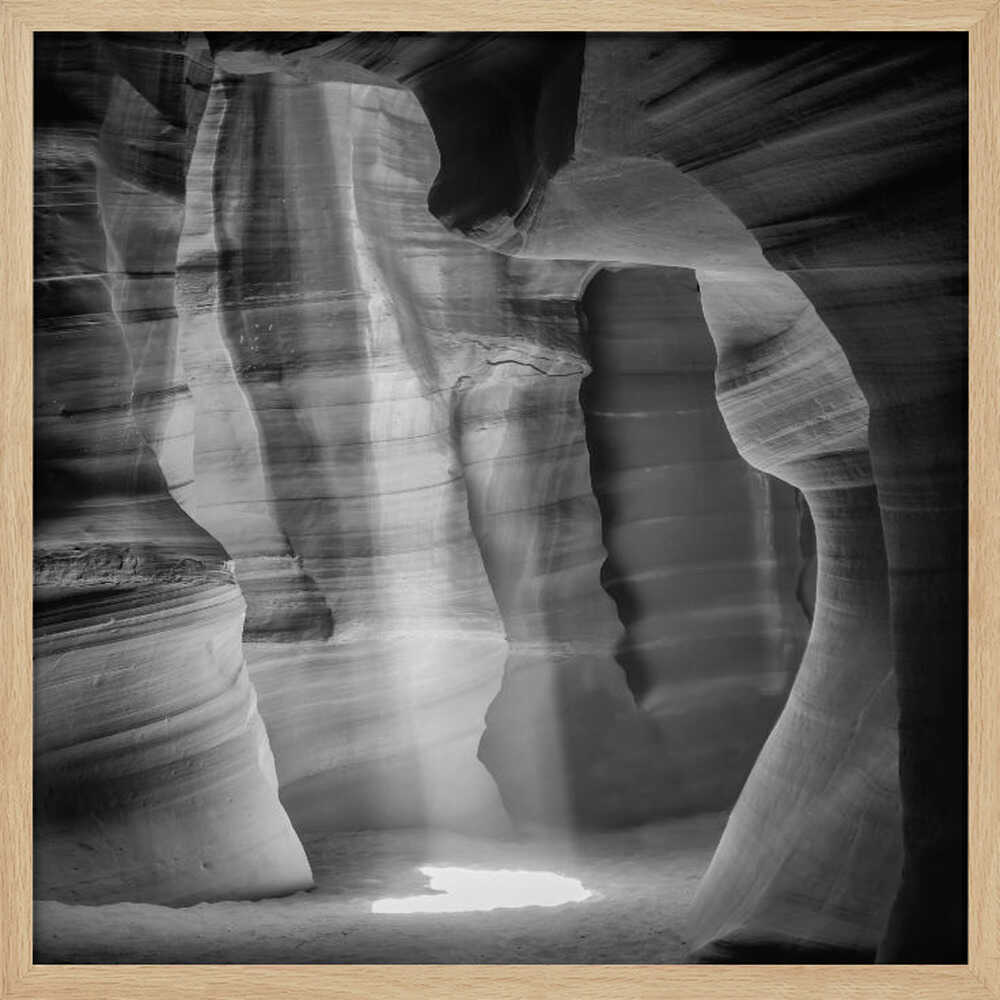 A dramatic black and white photo of a sunbeam lighting the sandy floor of a smooth, curved slot canyon.