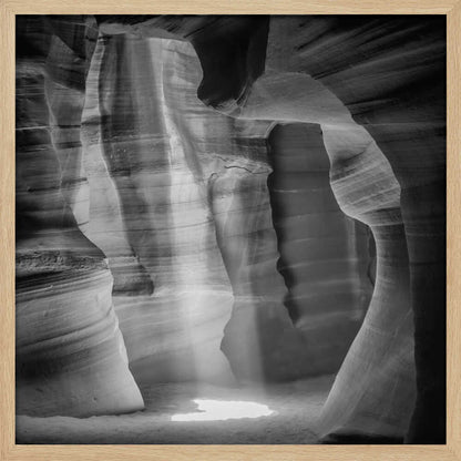 A dramatic black and white photo of a sunbeam lighting the sandy floor of a smooth, curved slot canyon.