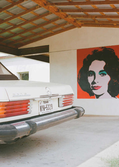A film-style photograph showing the rear of a classic white Mercedes-Benz parked under a carport with exposed wooden beams. On the adjacent white wall hangs a vibrant red pop-art portrait of a woman with dark hair. Artwork