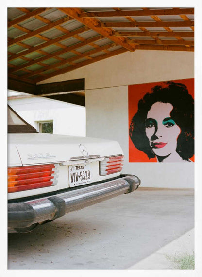 A film-style photograph showing the rear of a classic white Mercedes-Benz parked under a carport with exposed wooden beams. On the adjacent white wall hangs a vibrant red pop-art portrait of a woman with dark hair. Artwork
