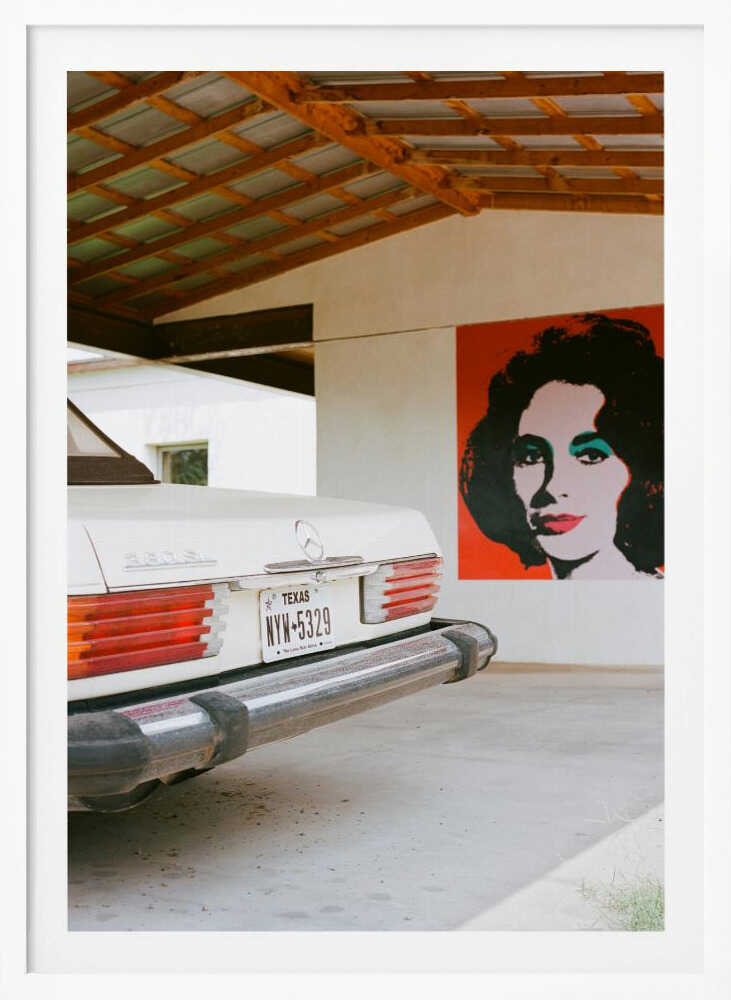 A film-style photograph showing the rear of a classic white Mercedes-Benz parked under a carport with exposed wooden beams. On the adjacent white wall hangs a vibrant red pop-art portrait of a woman with dark hair. Artwork