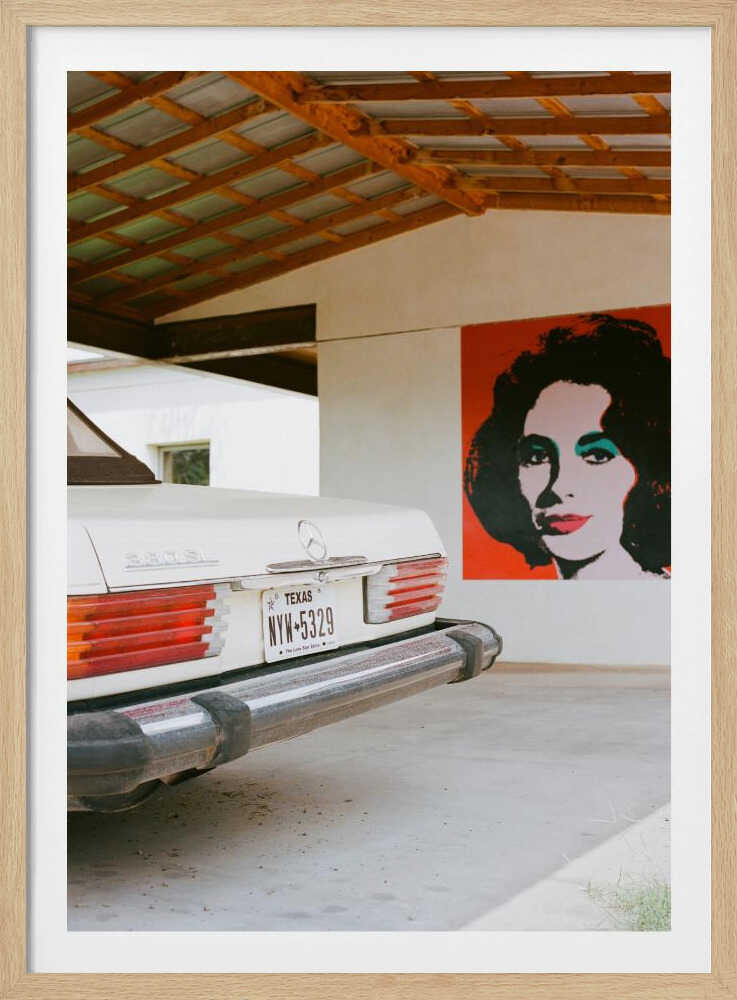 A film-style photograph showing the rear of a classic white Mercedes-Benz parked under a carport with exposed wooden beams. On the adjacent white wall hangs a vibrant red pop-art portrait of a woman with dark hair. Artwork