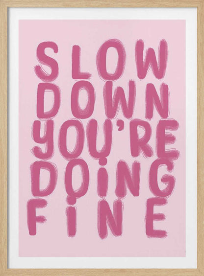 A framed art print featuring the phrase 'SLOW DOWN YOU'RE DOING FINE' in a bold, dark pink, brush-stroke font. The words are stacked vertically against a light pink background, all enclosed within a simple black frame. Artwork
