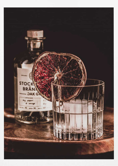 A moody still life photograph of a cocktail in a cut-crystal glass filled with ice. A dried blood orange slice garnishes the rim. In the soft-focus background, a bottle of gin rests on a dark wooden tray against a black backdrop. Wall Art