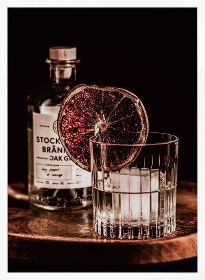A moody still life photograph of a cocktail in a cut-crystal glass filled with ice. A dried blood orange slice garnishes the rim. In the soft-focus background, a bottle of gin rests on a dark wooden tray against a black backdrop. Wall Art