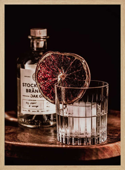 A moody still life photograph of a cocktail in a cut-crystal glass filled with ice. A dried blood orange slice garnishes the rim. In the soft-focus background, a bottle of gin rests on a dark wooden tray against a black backdrop. Wall Art