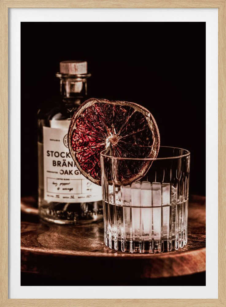 A moody still life photograph of a cocktail in a cut-crystal glass filled with ice. A dried blood orange slice garnishes the rim. In the soft-focus background, a bottle of gin rests on a dark wooden tray against a black backdrop. Wall Art