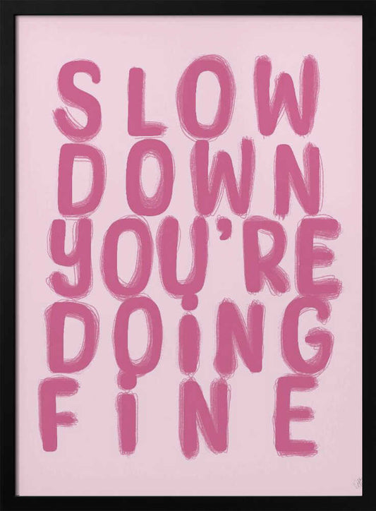 A framed art print featuring the phrase 'SLOW DOWN YOU'RE DOING FINE' in a bold, dark pink, brush-stroke font. The words are stacked vertically against a light pink background, all enclosed within a simple black frame. Artwork