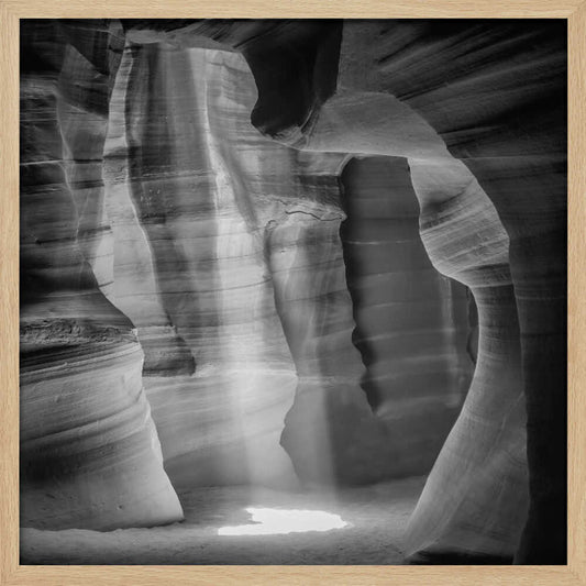 A dramatic black and white photo of a sunbeam lighting the sandy floor of a smooth, curved slot canyon.