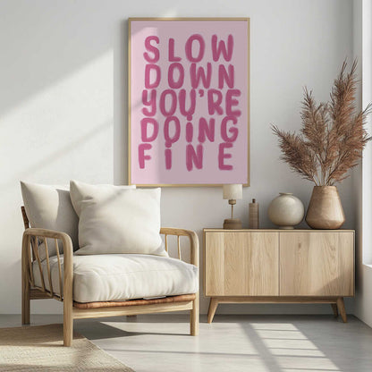 A framed art print featuring the phrase 'SLOW DOWN YOU'RE DOING FINE' in a bold, dark pink, brush-stroke font. The words are stacked vertically against a light pink background, all enclosed within a simple black frame. Artwork
