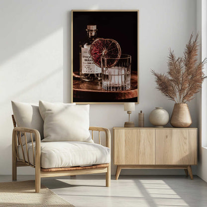 A moody still life photograph of a cocktail in a cut-crystal glass filled with ice. A dried blood orange slice garnishes the rim. In the soft-focus background, a bottle of gin rests on a dark wooden tray against a black backdrop. Wall Art